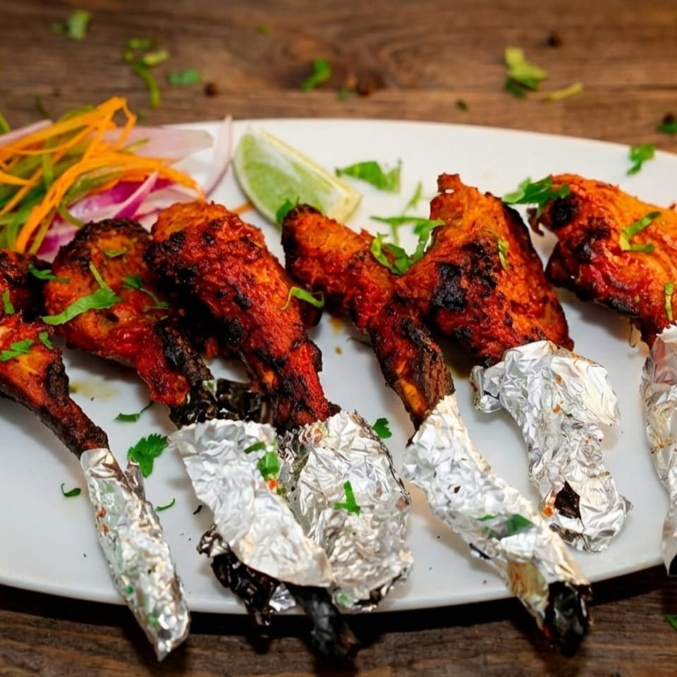A plate of Indian chicken lollipops with foil-wrapped bones, garnished with lime, onions, and cilantro.
