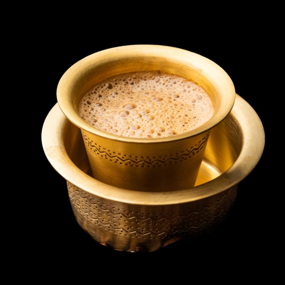 Frothy South Indian filter coffee in a traditional brass dabara set against a black background.