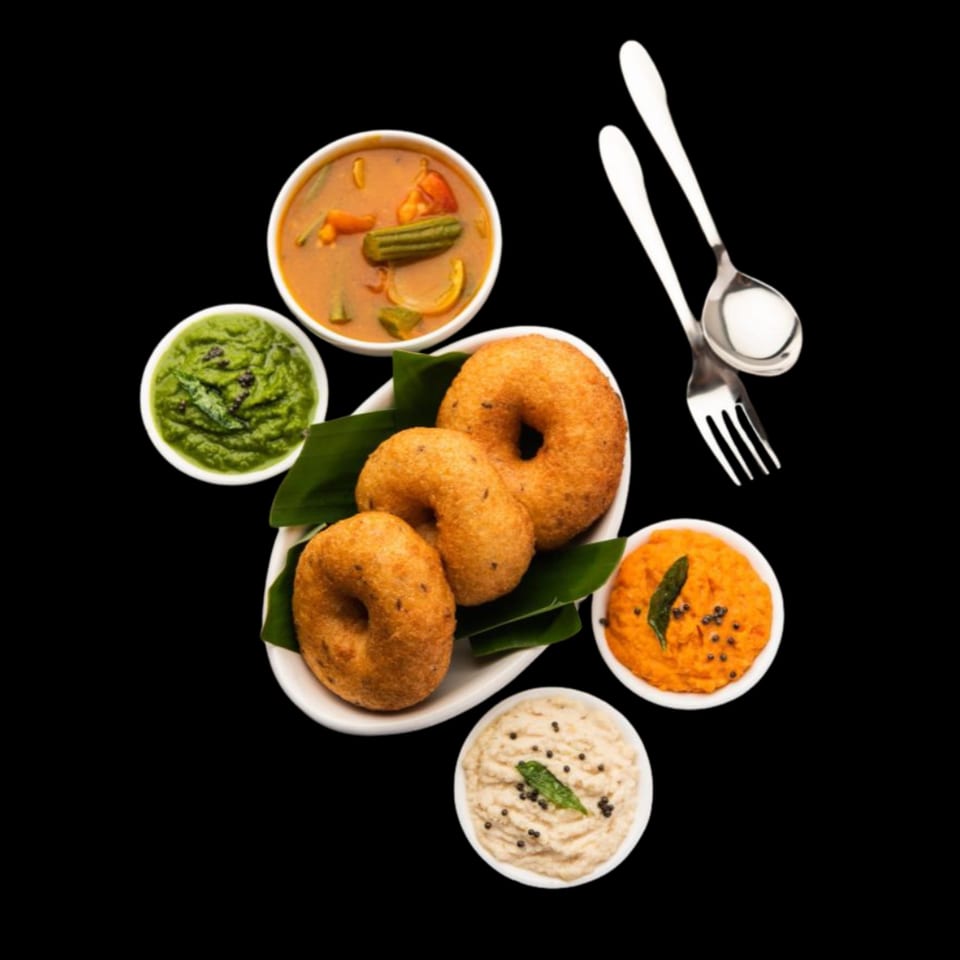 Three golden-brown medu vada on banana leaves, surrounded by bowls of sambar and three chutneys.