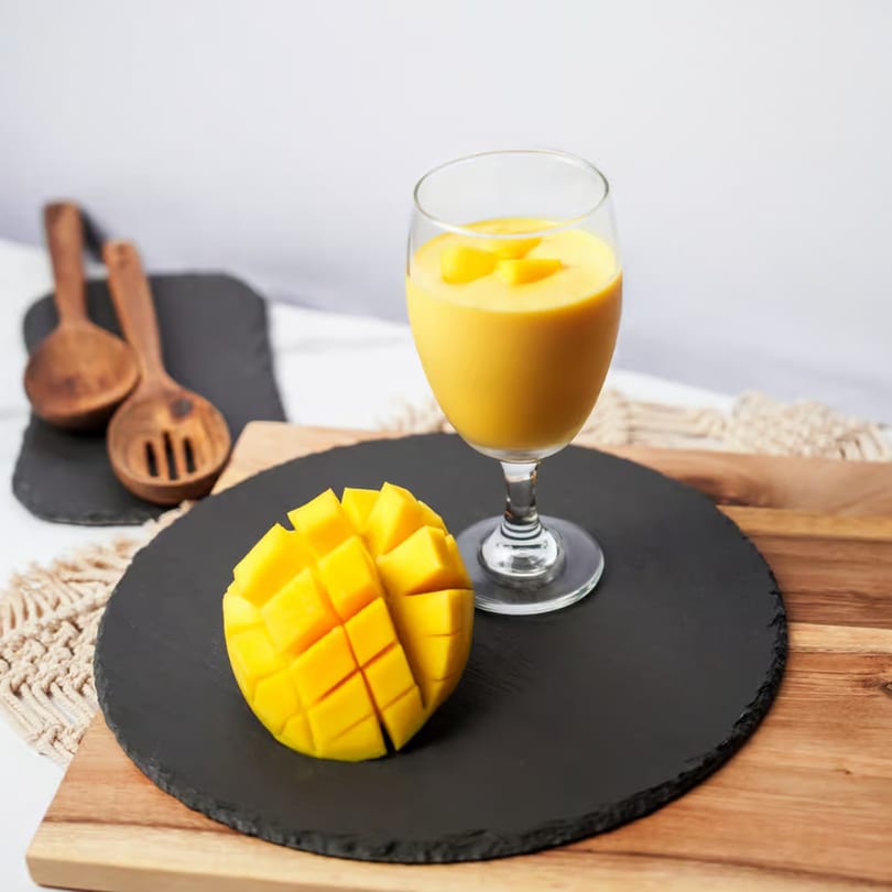 A glass of mango lassi with diced mango on a black slate and wooden board.