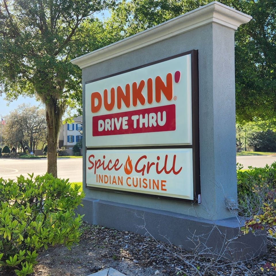 Outdoor sign for Spice Grill Indian Cuisine and Dunkin' Drive Thru, surrounded by green bushes and trees.