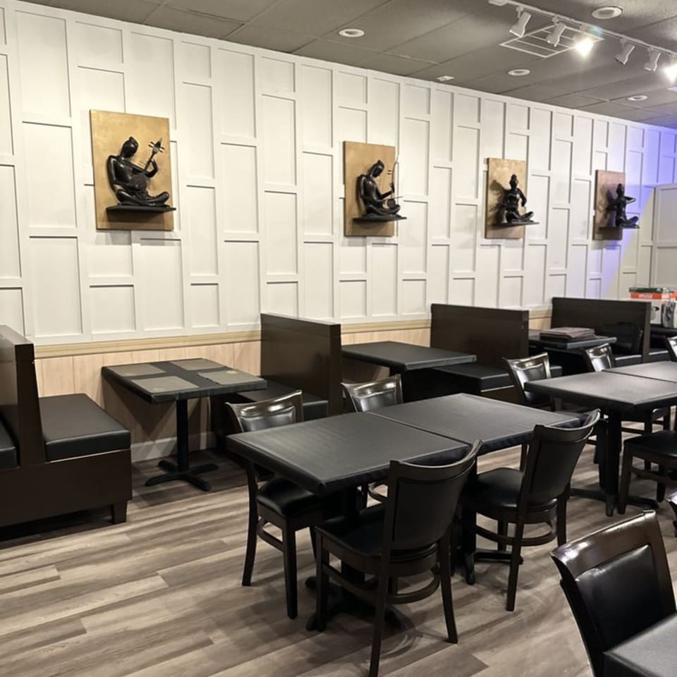 Empty restaurant dining room with dark tables, chairs, booths, and wall art on a light paneled wall.