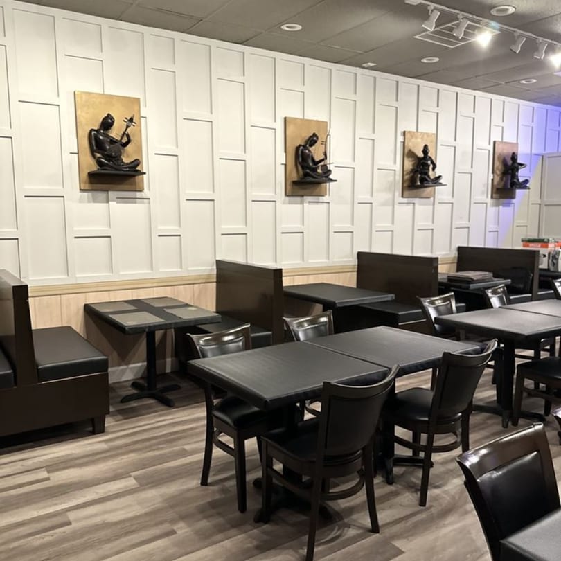 Empty restaurant dining room with dark tables, chairs, booths, and wall art on a light paneled wall.